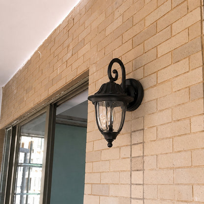 Black metal outdoor wall light lifestyle image with clear glass shade mounted on a tan brick entryway patio wall.