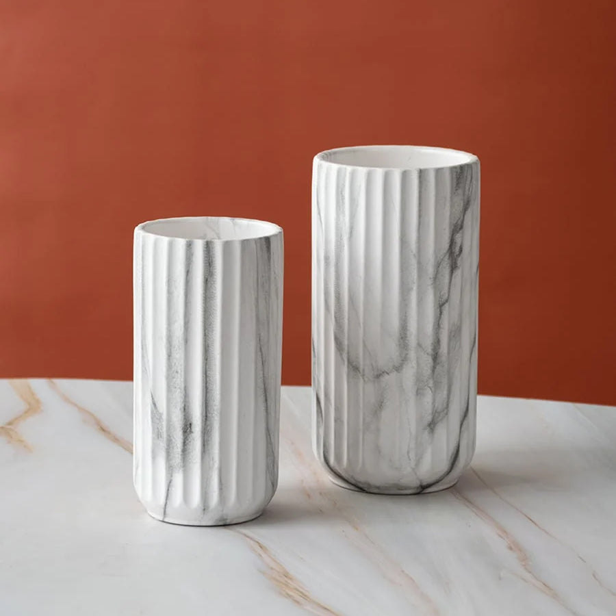 Two white fluted ceramic vases with gray marble veining, displayed on a white marble surface against a terracotta orange background.