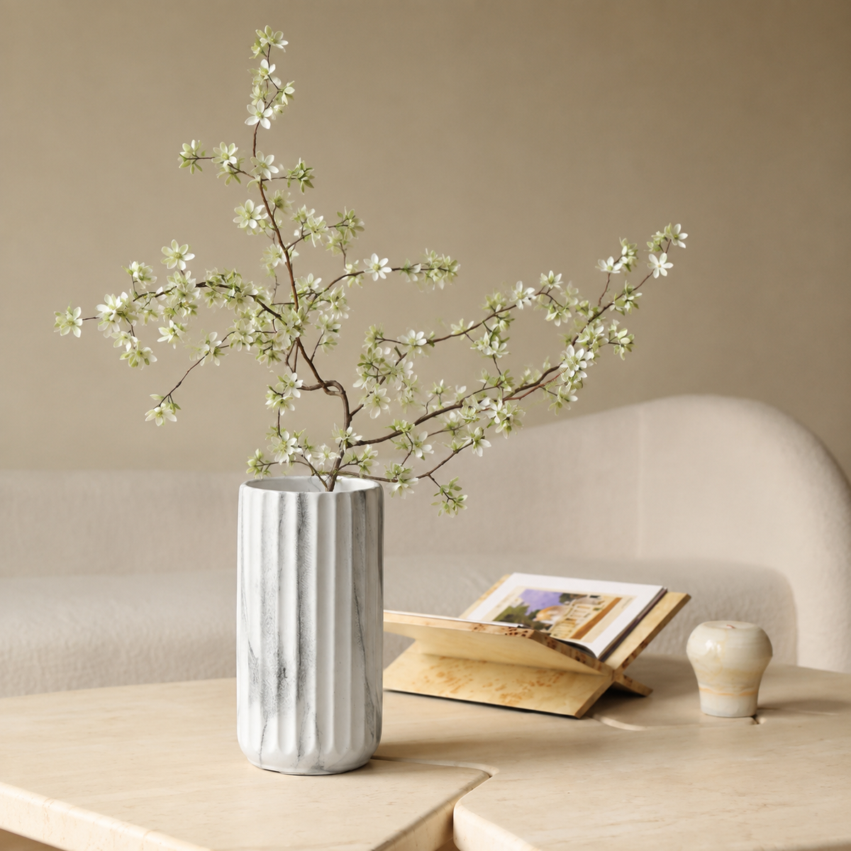 A ribbed white ceramic vase filled with delicate white blossoms on a light wood table, styled in a cozy living room setting with a sofa, an open book, and a small decorative pot.