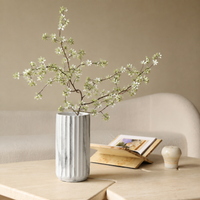 A ribbed white ceramic vase filled with delicate white blossoms on a light wood table, styled in a cozy living room setting with a sofa, an open book, and a small decorative pot.