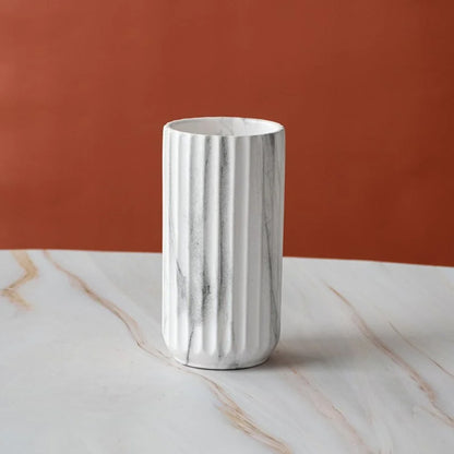 A white fluted ceramic vase with gray marble veining, standing on a white marble surface against a terracotta orange background.