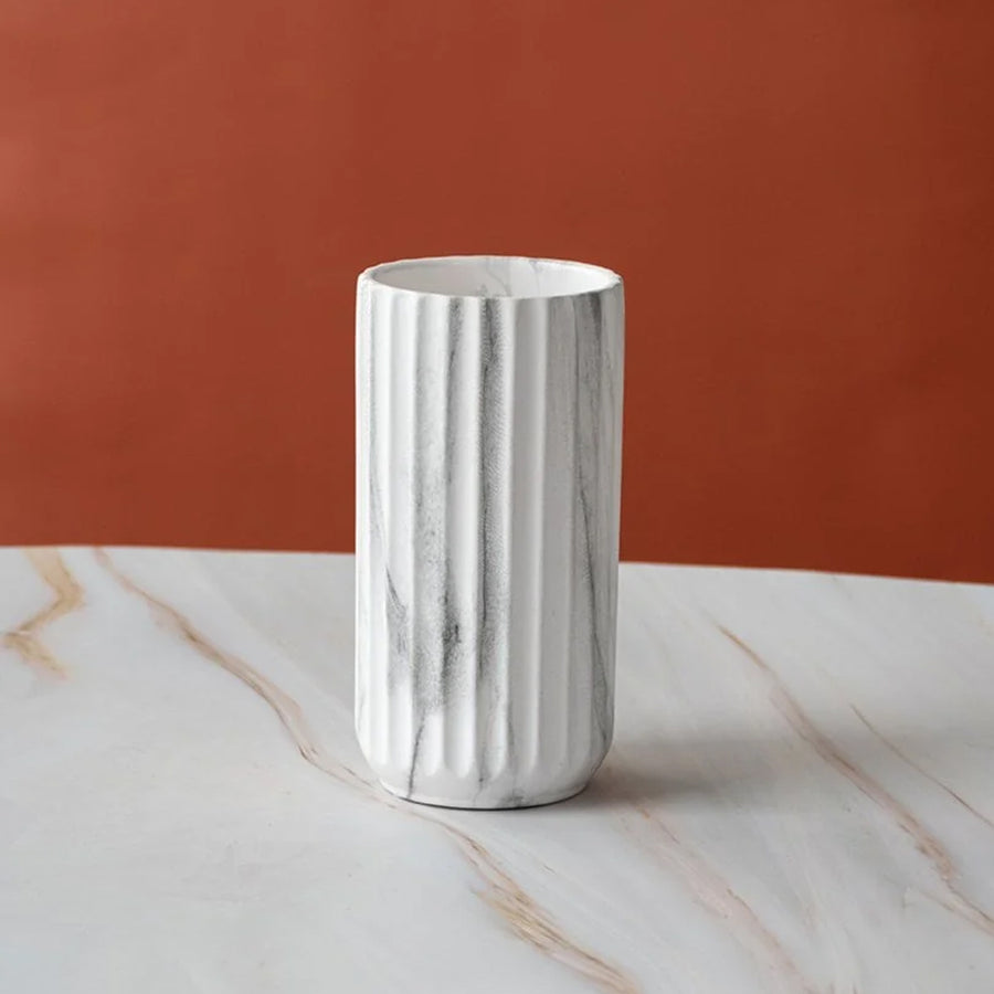 A white fluted ceramic vase with gray marble veining, standing on a white marble surface against a terracotta orange background.
