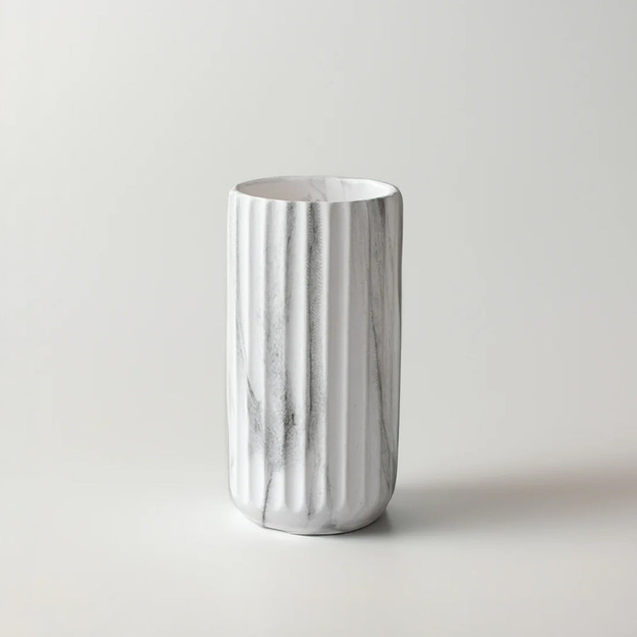 A white ceramic cylindrical cup or tumbler with vertical fluted ridges and a subtle gray marble pattern, set against a plain light gray background.