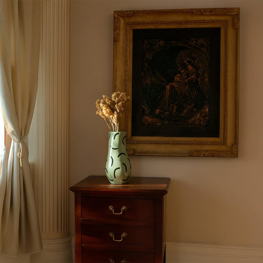 Green ceramic vase with black abstract patterns styled on a wooden chest with dried flowers adding artistic decor to a warm classic interior setting.