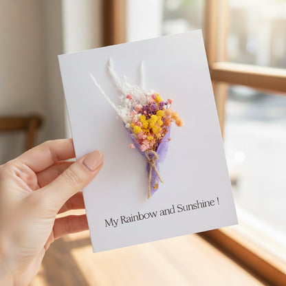 Hand holding a white greeting card with a mini purple and yellow dried flower bouquet in bright warm indoor sunlight.