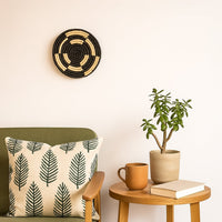 A handwoven natural and black rattan wall decor plate styled above a wooden side table with a potted plant and ceramic mug creating warm modern cozy home decor vibes.