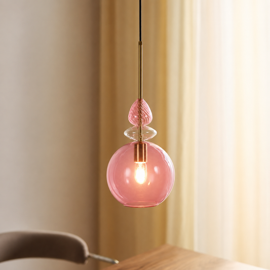 A pink glass pendant light hanging in a warm, softly lit interior space with curtains and a wooden table visible in the background.