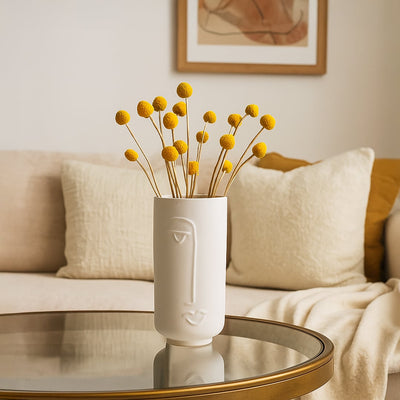 White ceramic face vase displayed on a glass coffee table in a cozy living room with soft neutral cushions and warm decor.