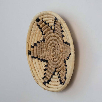 Handwoven seagrass round wall basket decor in natural beige and black with raised spiral star profile on white background.