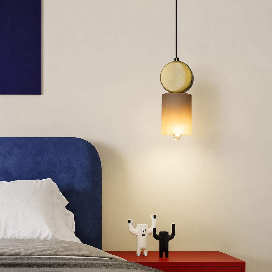 A modern pendant light with a round gold-toned top and a warm amber cylindrical shade hangs above a red bedside table in a bedroom setting.