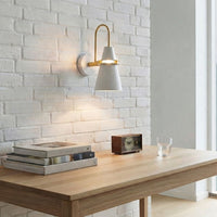 Contemporary white metal cone wall sconce with a curved brass arm mounted on a white painted brick wall above a wooden study desk.