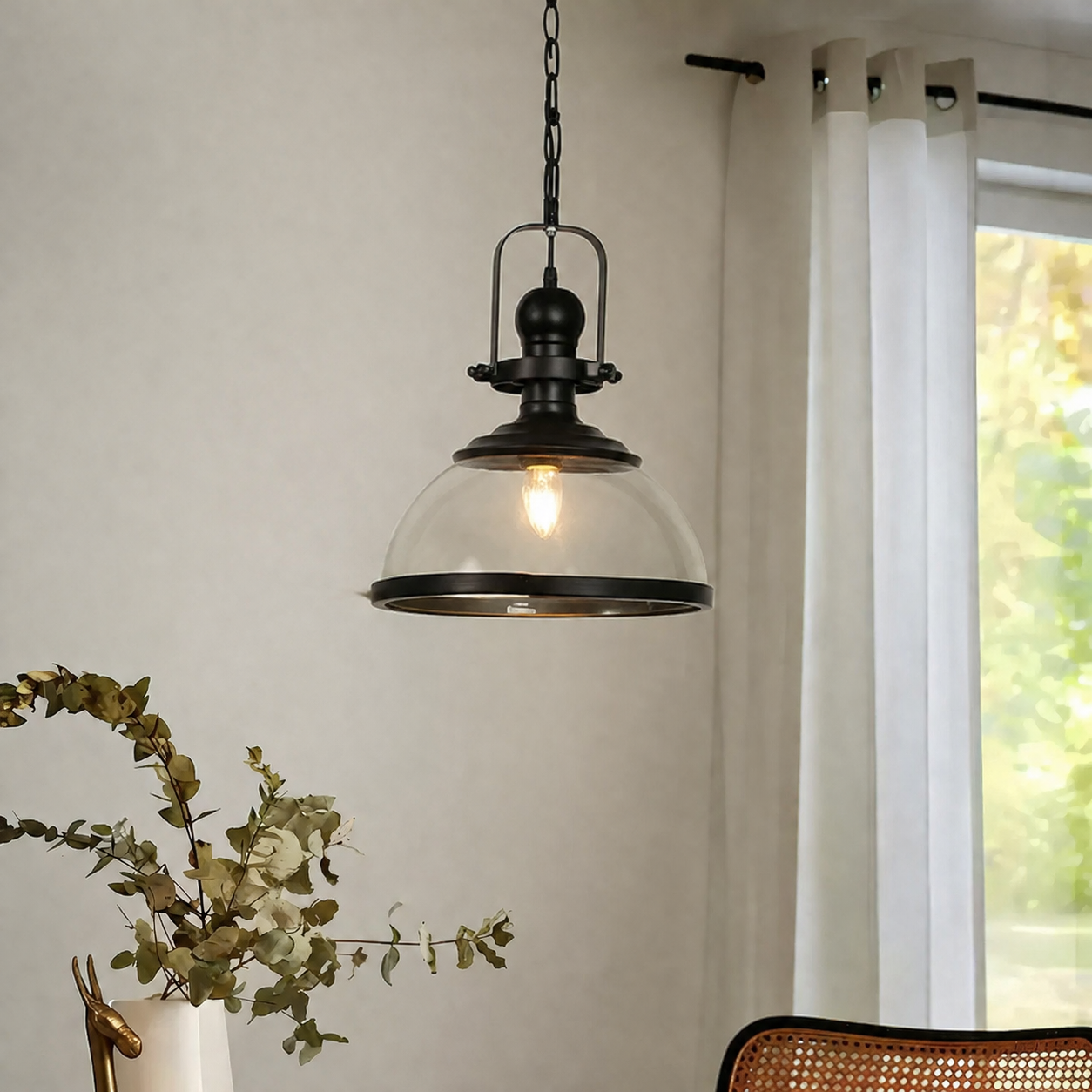 A hanging pendant light with a black metal frame and a clear glass dome shade, shown lit in a real interior setting with a window, curtain, and decorative plant visible.