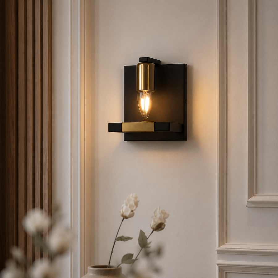 A modern wall sconce light fixture with a matte black rectangular wall mount, a brushed gold cylindrical shade, and a glowing warm Edison bulb, mounted on a cream-colored paneled wall.