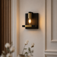 A modern wall sconce light fixture with a matte black rectangular wall mount, a brushed gold cylindrical shade, and a glowing warm Edison bulb, mounted on a cream-colored paneled wall.