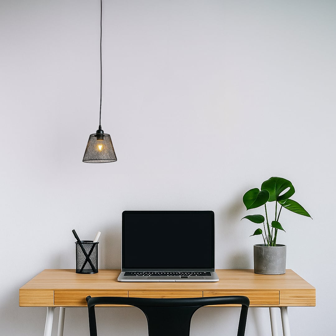 Modern black mesh pendant light hanging above a minimalist wooden desk with a laptop and plant creating a clean work setting with warm ambient glow.