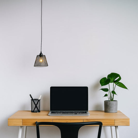 Modern black mesh pendant light hanging above a minimalist wooden desk with a laptop and plant creating a clean work setting with warm ambient glow.