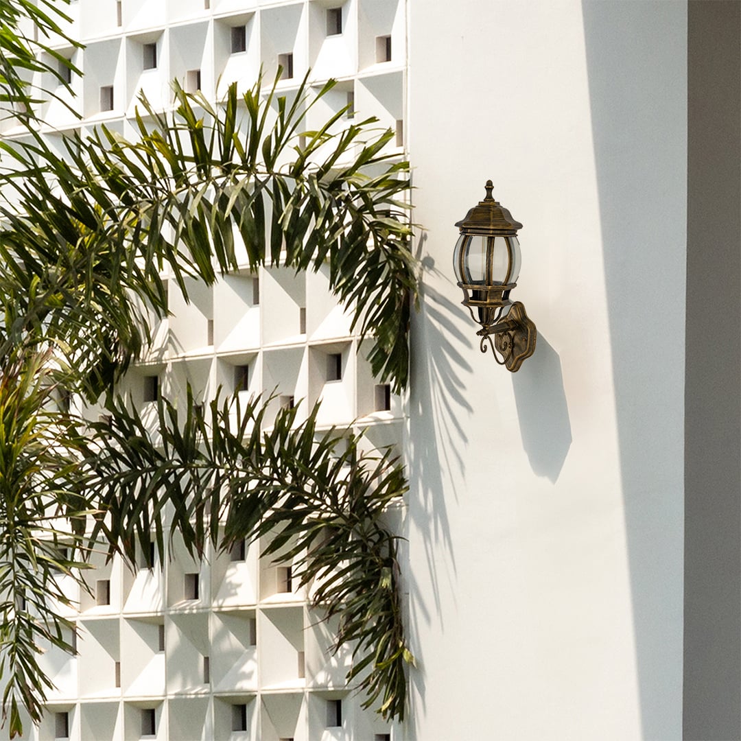 Antique gold traditional wall lamp with clear glass mounted on white decorative breeze block wall beside palm trees.