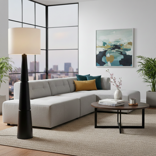 Modern black floor lamp with tall tapered base and white drum shade glowing beside sectional sofa in bright city living room interior style.