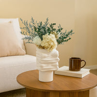 White sculptural ceramic vase with modern textured design styled on a wooden coffee table with flowers beside a sofa in a cozy neutral living room.