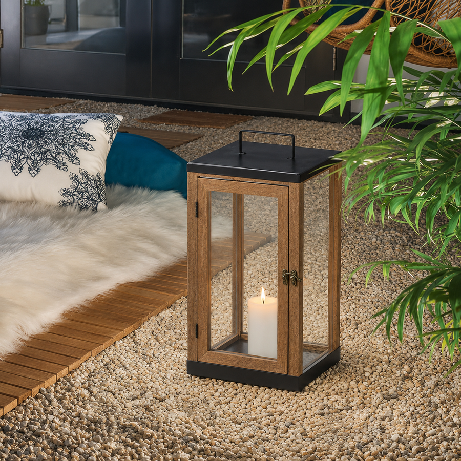 A wooden lantern candle holder with a black metal top and base, containing a lit white candle, placed on a pebble surface in a styled indoor setting with a rug, pillows, and green plant leaves.
