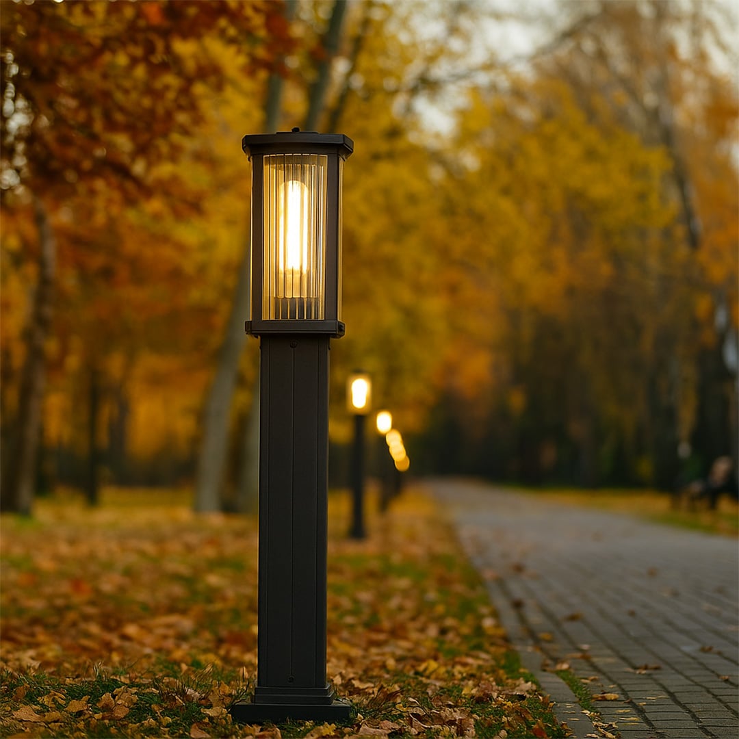 Black outdoor bollard light with ribbed glass glowing warmly along a tree lined pathway creating a cozy inviting atmosphere in an autumn garden setting.