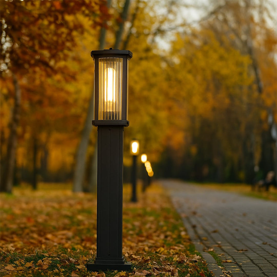 Black outdoor bollard light with ribbed glass glowing warmly along a tree lined pathway creating a cozy inviting atmosphere in an autumn garden setting.