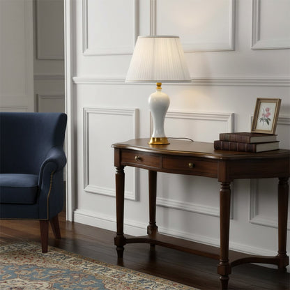 Table lamp with white ceramic base gold detailing and pleated shade placed on a wooden console adding warm ambient lighting to elegant living room decor.