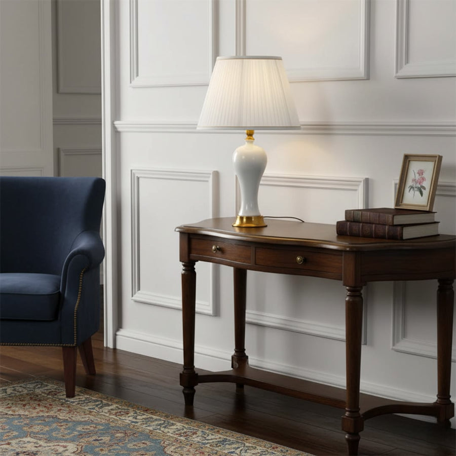 Table lamp with white ceramic base gold detailing and pleated shade placed on a wooden console adding warm ambient lighting to elegant living room decor.