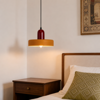 A modern pendant light with a dark red cylindrical shade and a wide warm yellow-gold drum shade, hanging in a bedroom next to a wooden nightstand and bed.
