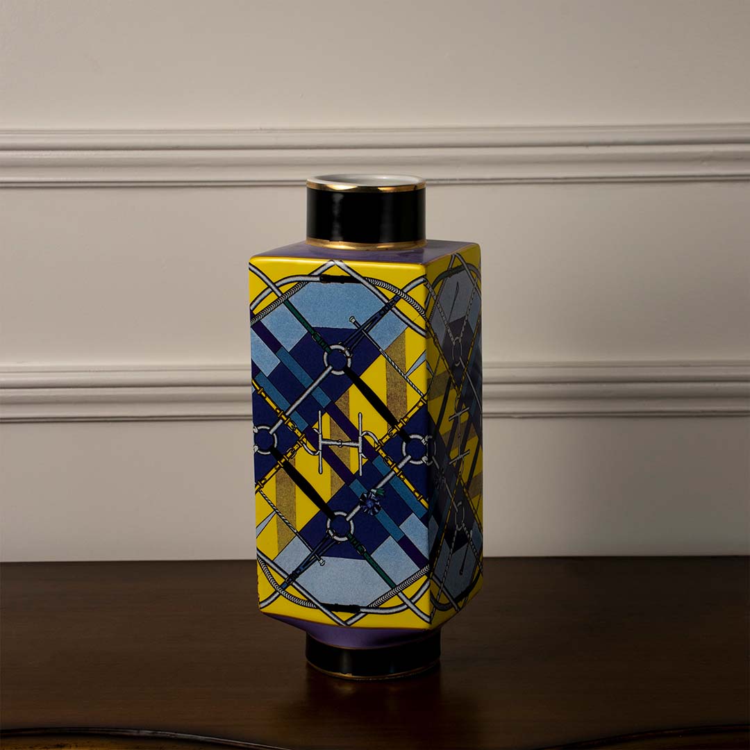 Decorative ceramic vase with blue and yellow geometric pattern and black top placed on wooden table against neutral wall background for modern home decor styling.