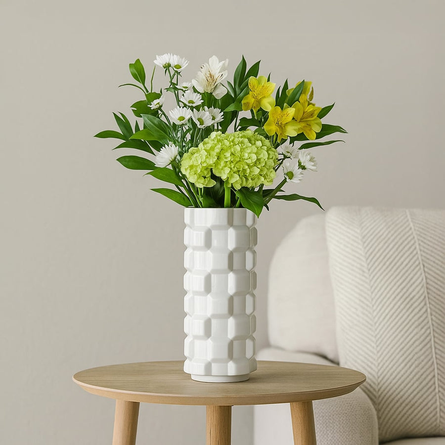 White ceramic flower vase with green foliage placed on round wooden table near sofa creating fresh and elegant home decor style.