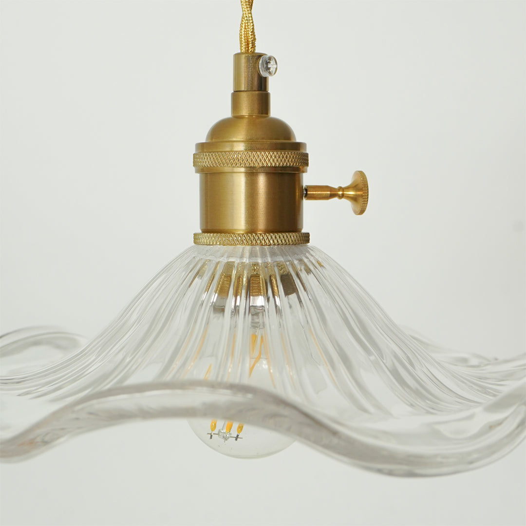 A close-up of a hanging light fixture with a brushed brass metal socket and cord, and a clear ribbed glass shade shaped like a wavy octopus tentacle, against a plain white background.