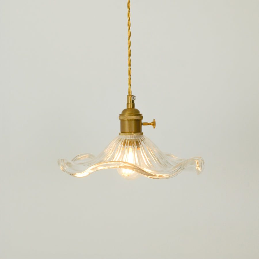 A vintage-style pendant light with a clear ribbed glass shade that has wavy, flared edges and a brass metal fixture, suspended from a brass cord against a light gray background.