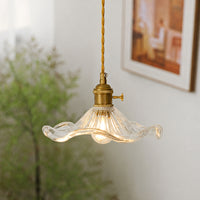 A hanging pendant light with a wavy clear glass shade and a brass-toned socket and cord, shown lit in a softly blurred indoor setting.