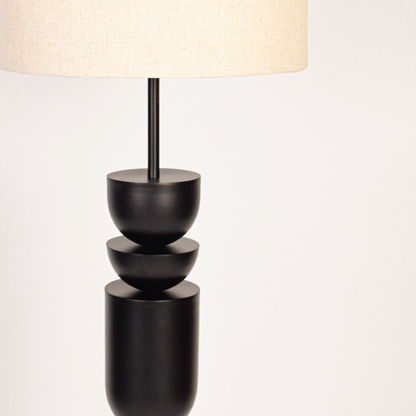 Black floor lamp on white background showing sculptural stacked base and tall fabric shade creating soft ambient glow.