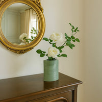 Green ceramic vase with ribbed texture holding white roses placed on a wooden console beneath a gold-framed mirror for an elegant decor setting.