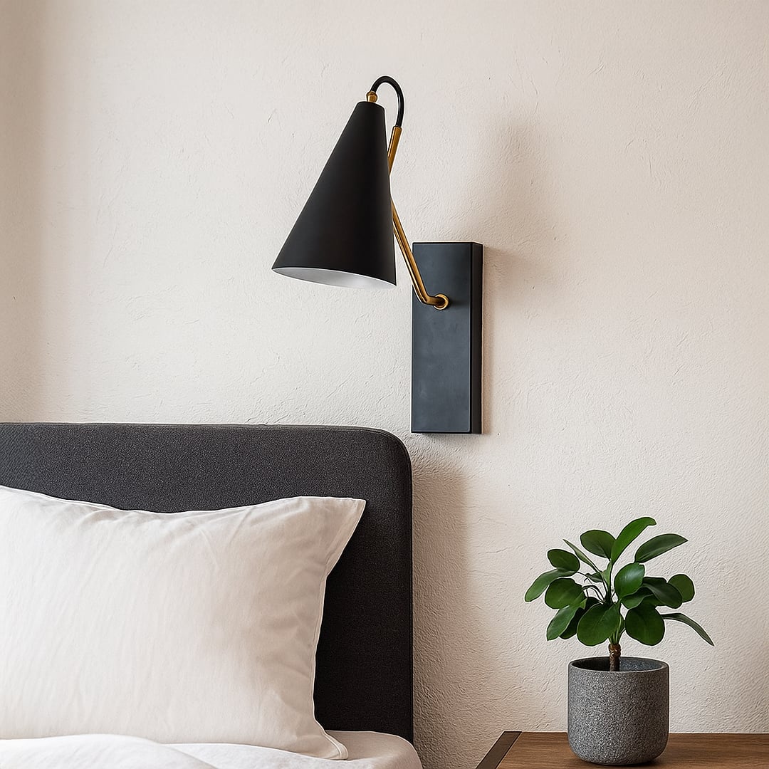 Black wall light with conical shade and gold arm mounted above bedside table with plant styled in modern bedroom interior.