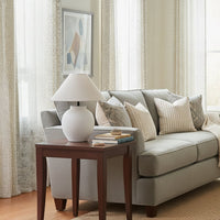 Lifestyle image of white ceramic table lamp with fabric shade placed on wooden side table beside sofa adding soft ambient living room lighting.