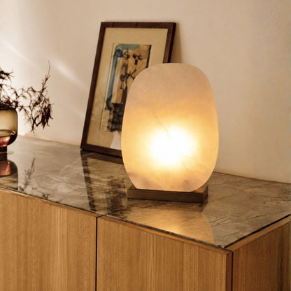 A glowing oval table lamp made of translucent stone sits on a wooden sideboard with a marble top, next to a framed picture and a small vase.