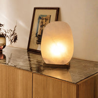 A glowing oval table lamp made of translucent stone sits on a wooden sideboard with a marble top, next to a framed picture and a small vase.
