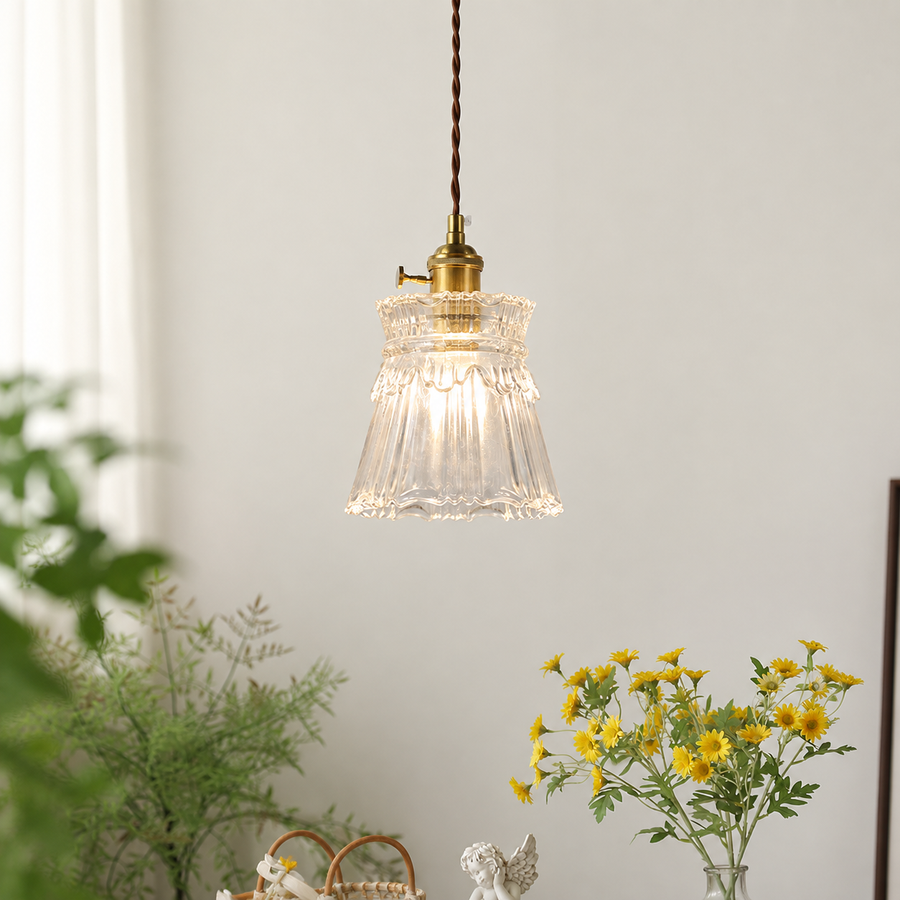 A vintage-style pendant light with a clear ribbed glass bell shade and a brass metal fitting, hanging from a brown cord in a softly lit room with plants and yellow flowers in the foreground.