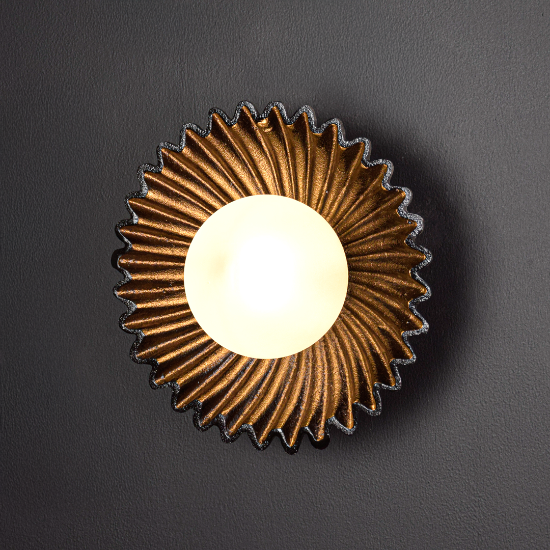 Wall light illuminated on a black background creating a dramatic warm halo that highlights the sculpted metal form and elegant modern appeal.