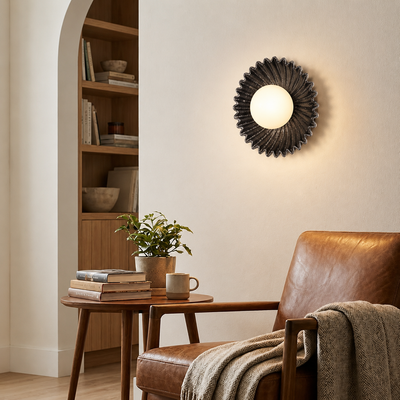 A round wall sconce with a dark textured sunburst frame and glowing warm light, mounted on a cream wall in a styled living room with a leather chair, side table, books, and a plant.