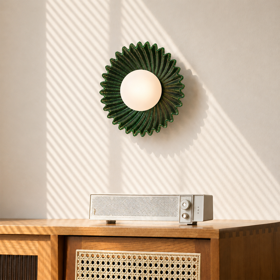 A green sunburst-shaped wall sconce with a white globe bulb mounted on a beige wall, casting diagonal shadows. Below the light, a silver and white radio sits on a wooden cabinet with a woven cane panel.