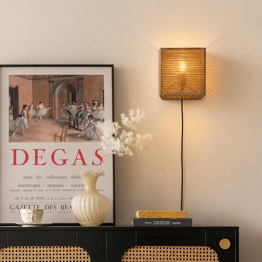A warm, square woven wall sconce lamp glowing with a yellow light, mounted on a beige wall above a black console table with a framed Degas poster, a cream vase, and books.