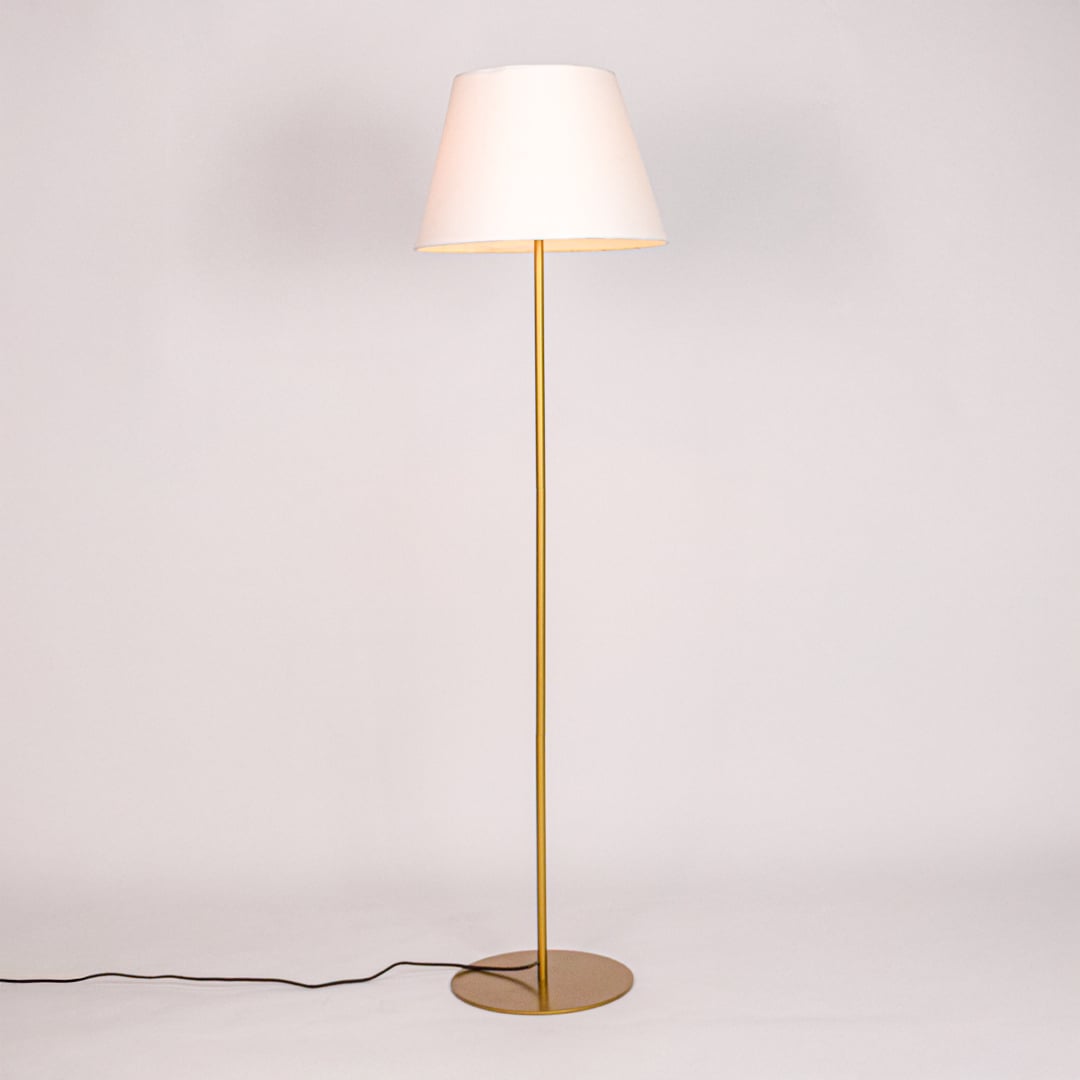 Gold floor lamp illuminated with warm glow through white fabric shade against minimal white studio background.