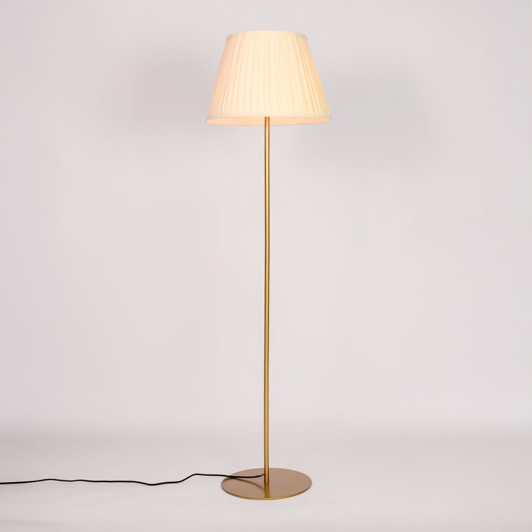 Gold metal floor lamp glowing warmly through pleated cream fabric shade on white background for cozy home Decor.