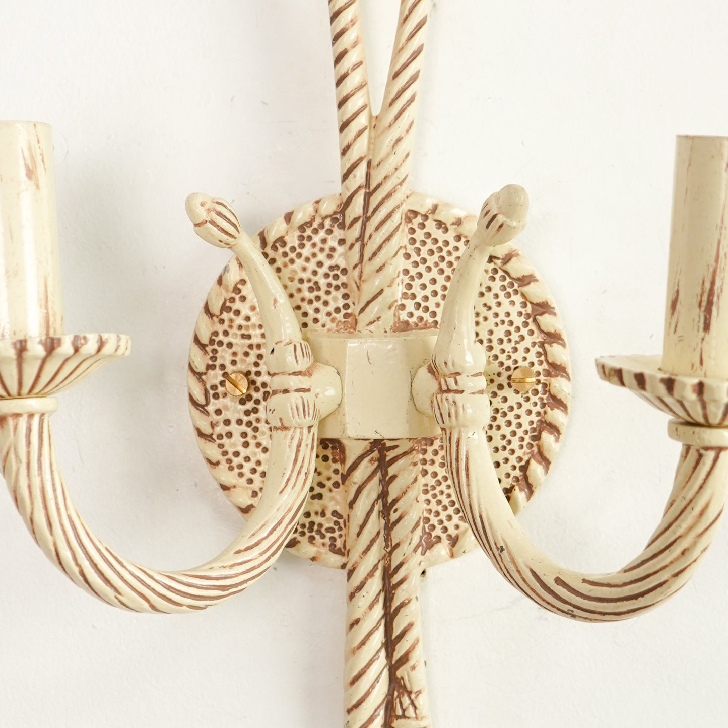 A close-up of a distressed cream and brown wall sconce with twisted rope-style arms and a circular patterned backplate.