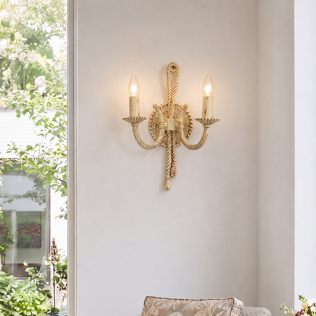 A two-light wall sconce with a gold-finished rope and lion motif, mounted on a light beige wall next to a window with green foliage visible outside.