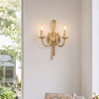 A two-light wall sconce with a gold-finished rope and lion motif, mounted on a light beige wall next to a window with green foliage visible outside.